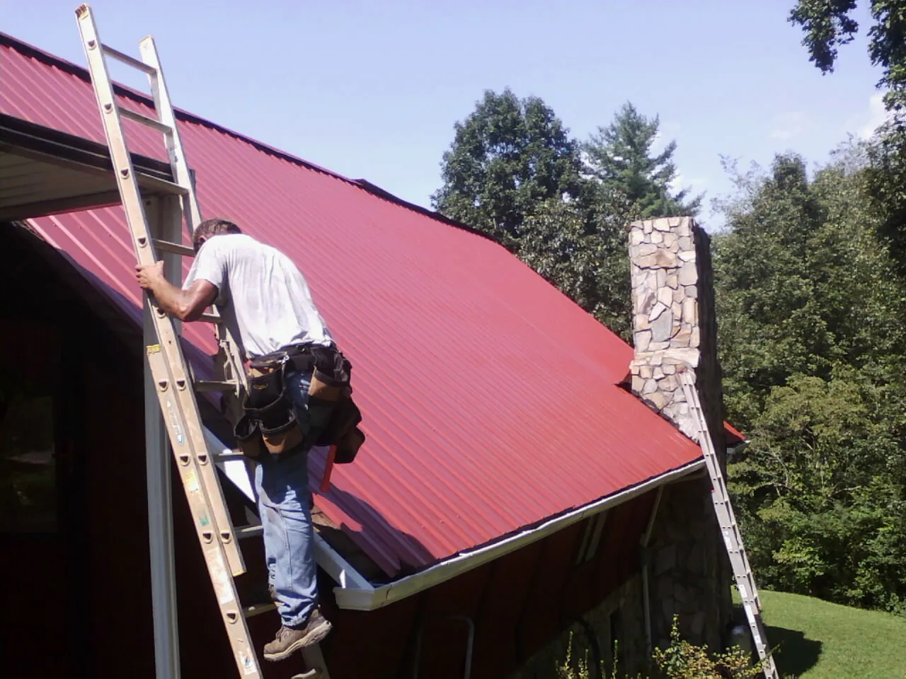 Roofer on a ladder beside a red metal roof and stone chimney — Soffit & Fascia Repair by Roofing Brothers in Twin Falls